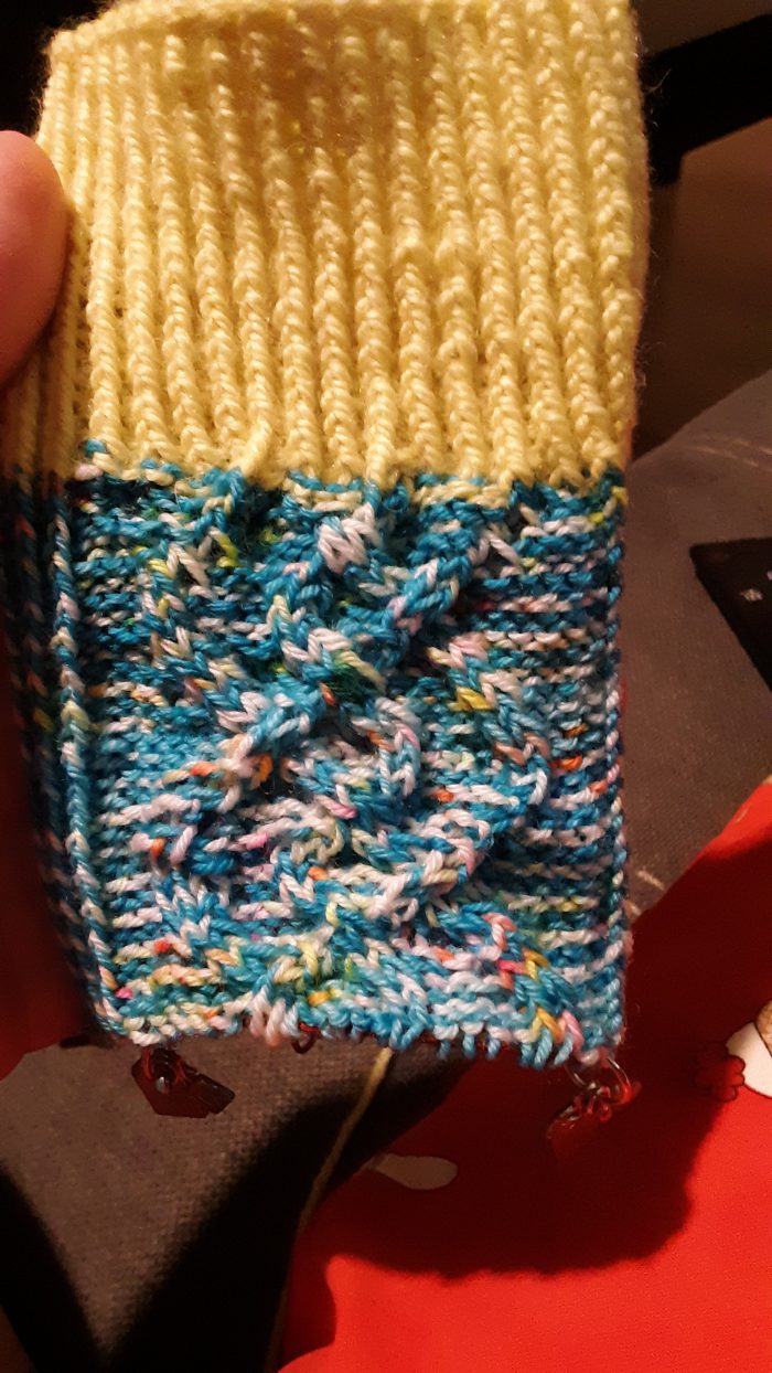 several inches of a knitted sock, with a yellow ribbed cuff and variegated blue leg knit in a cable pattern