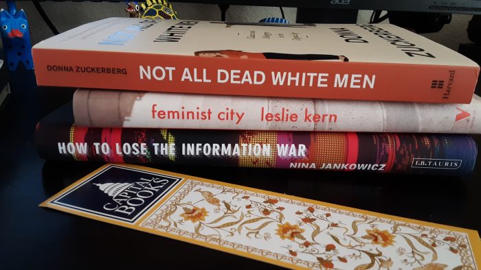 a stack of three hardcover books: Not All Dead White Men, Feminist City, and How to Lose the Information War