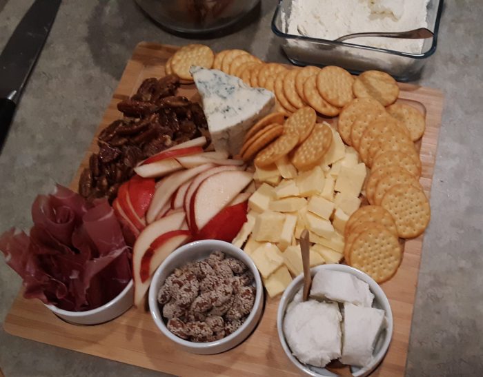 A wooden board covered in snacks> several kinds of cheese, nuts, crackers, proscuitto, and pear slices