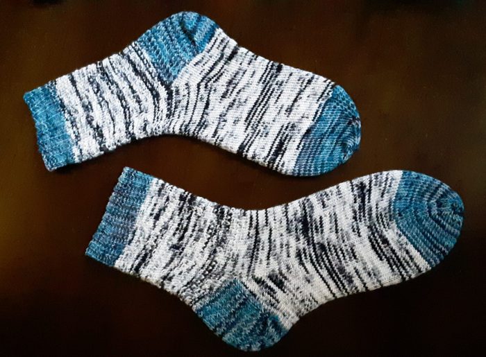 a pair of socks lying flat on a wood table. Main yarn is varigated black and white but the toe, heel, and cuff are a variegated blue