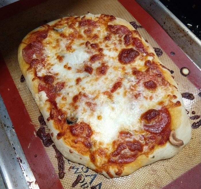 a homemade pizza on a rectangular sheet pan. Pepperoni, bubbly browned cheese, cashews and olives are on top