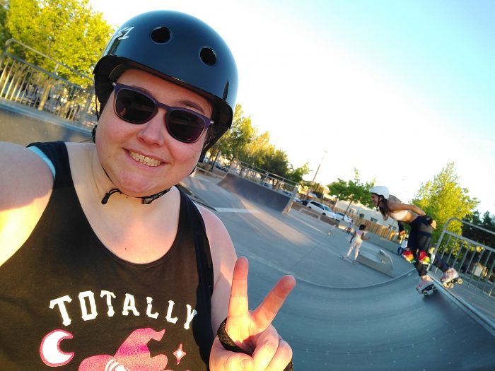 me, wearing a helmet and sunglasses, flashing a peace sign at teh skate park. A young woman on skates drops into a halfpipe behind me.
