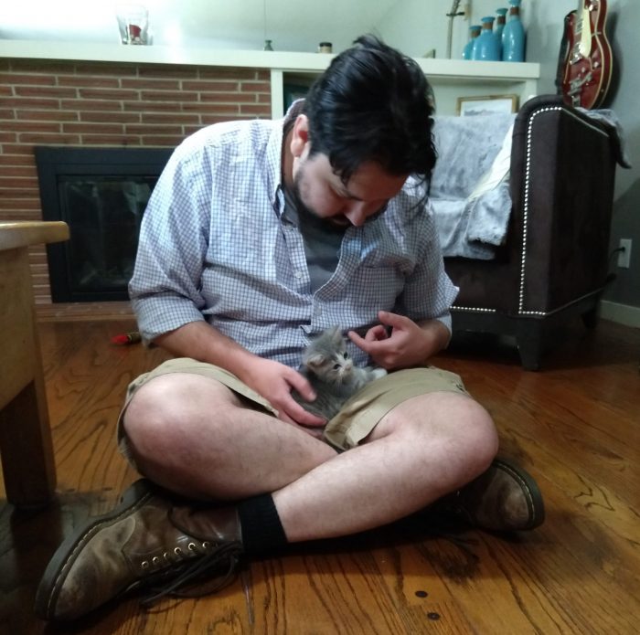 Kirk, sitting cross-legged on a hardwood floor, holding a tiny grey kitten in his lap