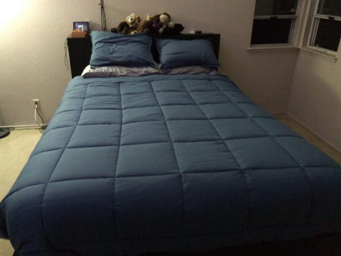 a light blue comforter over silver sheets, there are stuffed animals on the shelf above the bed