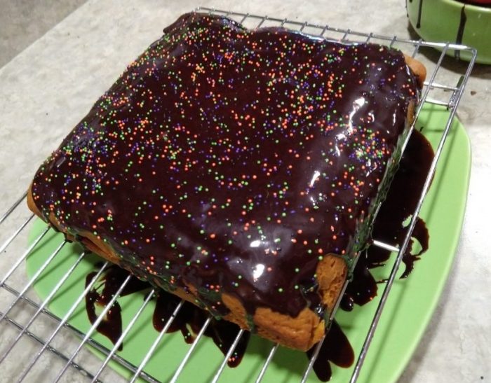 a square pumpkin cake on a wire rack, covered with chocolate glaze that is dripping off the cake, topped with orange, purple and green sprinkles