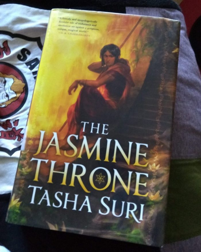 hardback book: The Jasmine Throne by Tasha Suri. Cover features a woman wearing a Sari crouched against a wall and looking out suspiciously
