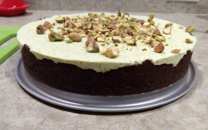 a tart sitting on the bottom part of a springform pan. You see an uneven chocolate crust up the side and a light green pistachio mousse topped with chopped pistachios