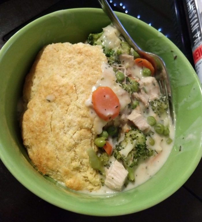 A bowl of turkey pot pie. The top of the pie is a hunk of biscuit and the inside has broccoli, peas, carrot, and turkey in an herbed, creamy sauce