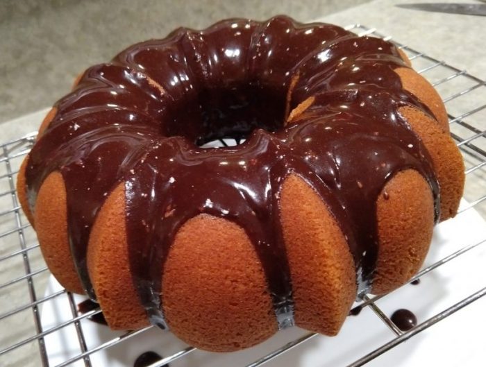 a bundt cake cooling on a wire rack. The cake is golden brown and there is a chocolate topping dripping down through the grooves of the cake