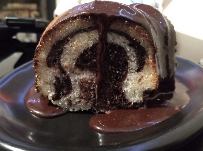 a slice of marbled bundt cake. The cake is vanilla with chocolate swirls. There's a chocolate sauce on top that's dripping down and pooling on the plate.