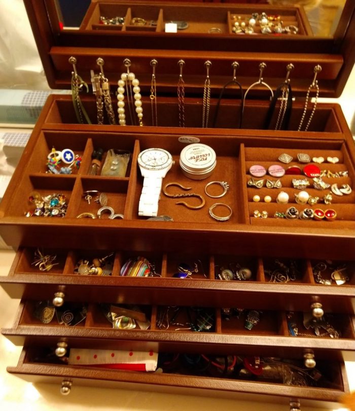 a four-tiered jewelery box with the lid open and each drawer slightly pulled out to reveal earrings (mostly). The inside of the lid has a mirror and there are hooks to hang necklaces.