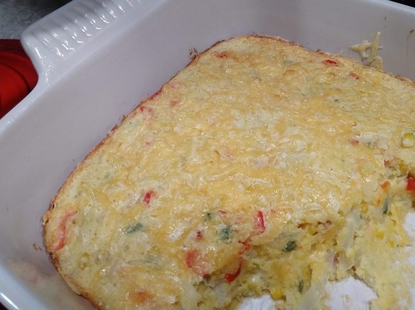 a casserole dish containing spoon bread—some already scooped out. The filling of onion, jalapeño, and red bell pepper is visible. There's cheese on top.