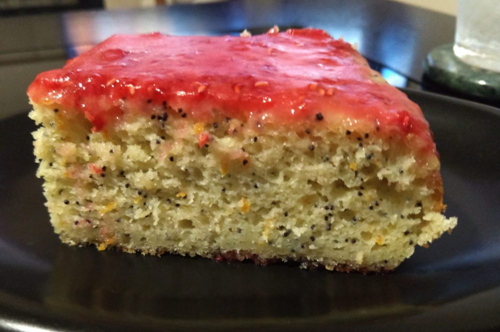 a rectangular slice of cake on a small plate. The cake is orange poppy seed cake with raspberry glaze.