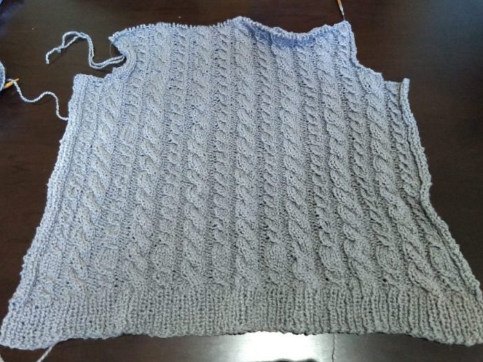 nearly finished back panel of a sweater. The yarn is grey and features a cabled design with alternating columns of cable styles all the way up the sweater.