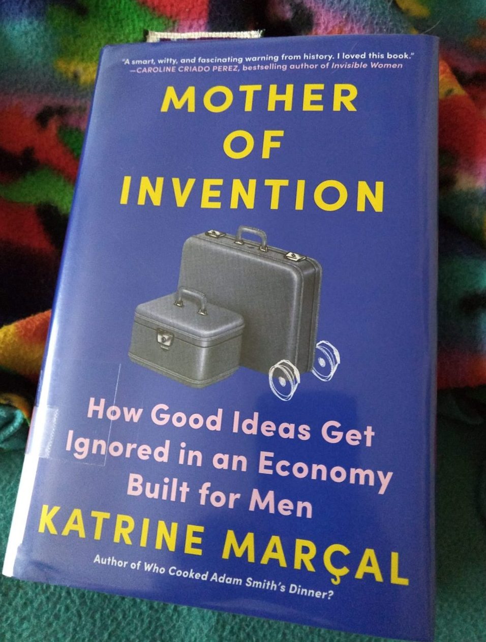 book: Mother of Invention: How Good Ideas Get Ignored in an Economy Built for Men. The cover is blue and minimalist, featuring a mid-century style suitcase