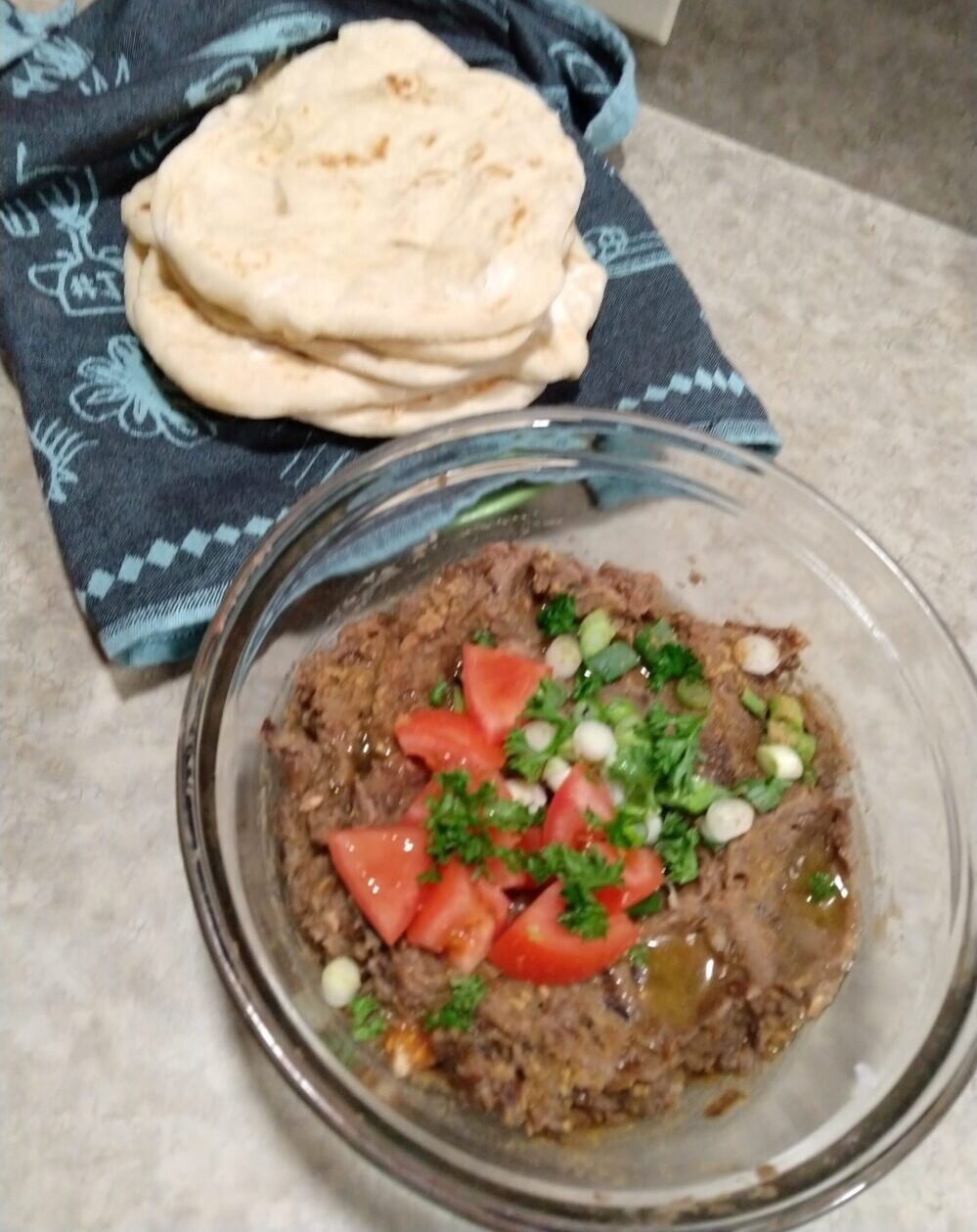 a slightly blurry photo of a bowl of bean dip topped with tomatoes, green onion, and parsley next to a stack of pita bread