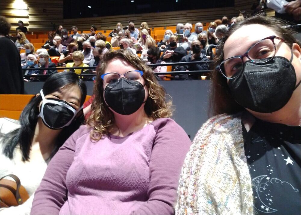 three women in a theater, masked and looking at the camera
