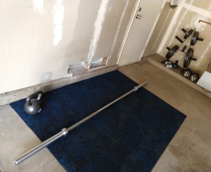 my garage with a cleared area covered in a small amount of rubber gym flooring. There is a weightlifting bar a nd a rack with a small set of weights