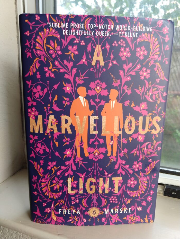 hardcover book: A Marvelous Light by Freyja Marske. The jacket design as a floral wallpaper-like pattern and there are silhouttes of two men