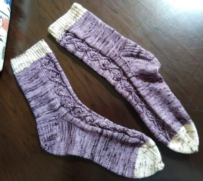 a pair of handknit socks with a varigated purple yarn as the main color and a creamy color on the cuff and toe. There's a small cable pattern running along the side of the sock.