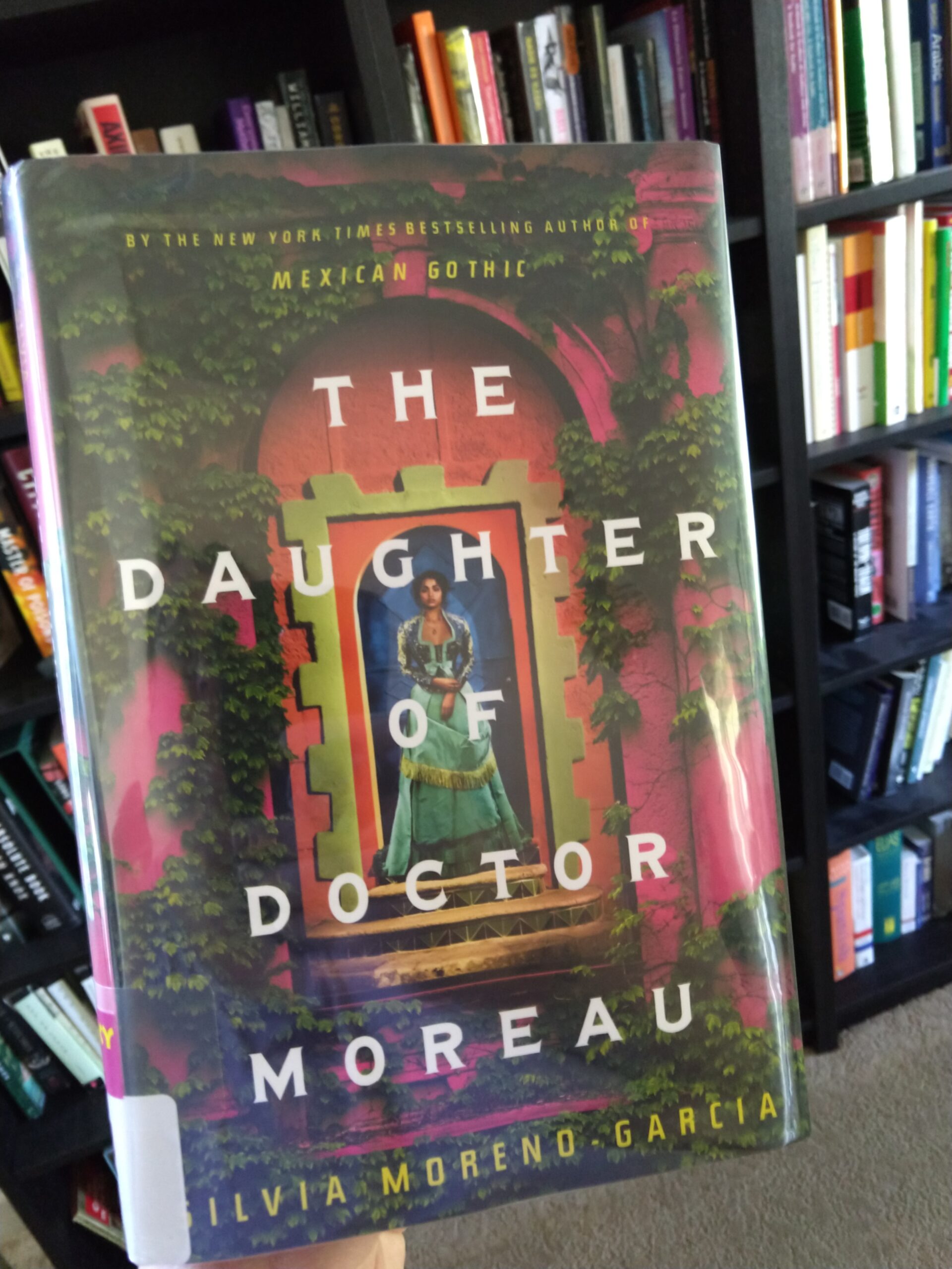 hardcover book from the library: The Daughter of Doctor Moreau by Silvia Moreno-Garcia. The cover shows a pretty young woman wearing a gown, standing in a doorway/arch that is covered in leaves