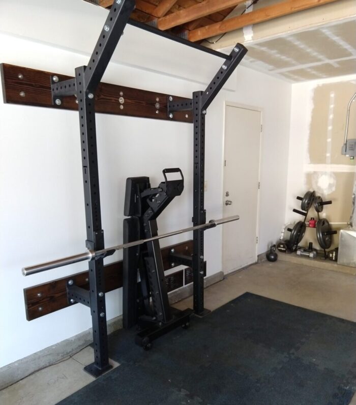 a view of our garage gym showing a squat rack that only stands about a foot from the wall and has a pull-up bar. There's a bench standing up and tucked in the rack. There are weight plates off to one side.