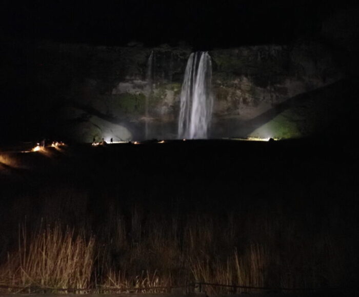 A waterfall seen at night. There are bright lights illuminating the waterfall and dimmer lights marking the walking path to it