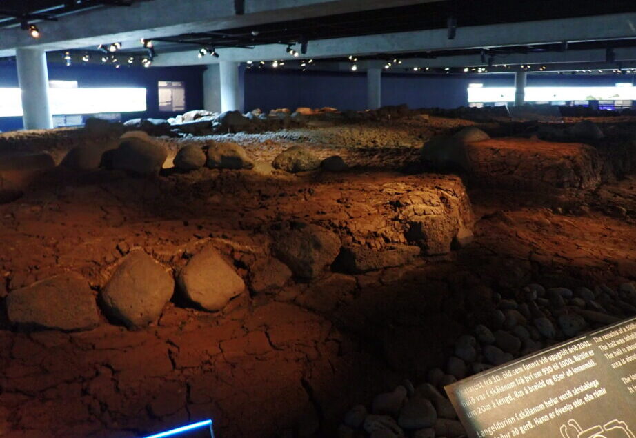 a museum exhibit below ground that displays the excavated remains of a place where people lived in the 10th century. Rocks and an outline of the dwelling remain.