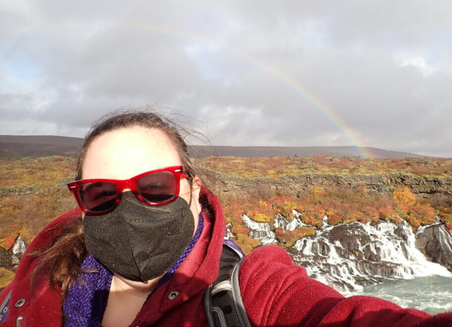 me, wearing extremely cool red sunglasses and a less cool but very safe facemask, in front of the Hraunfossar. The waterfalls look cool. The fall folliage is pretty and there's a wonderful rainbow in the background