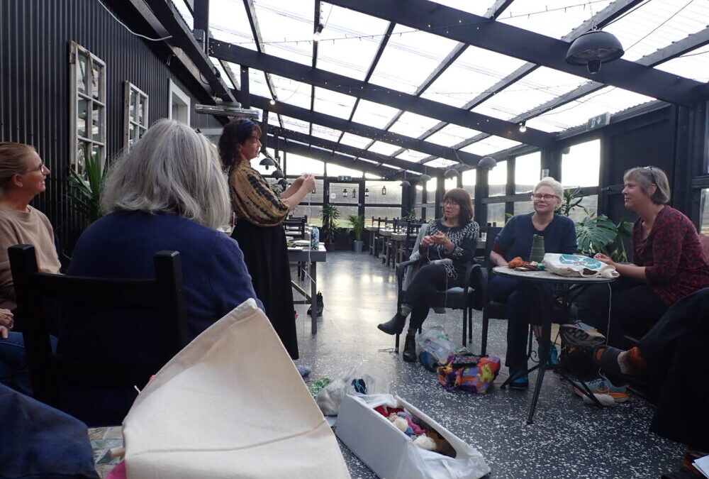 a sunroom in which a group of knitters are seated at small tables. Our instructor is demonstrating a knitting technique.