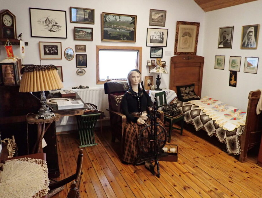 A room featuring a small bed, walls covered in pictures, a desk, and a mannequin of Halldora in traditional Icelandic costume seated at a spinning wheel
