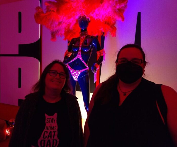 foreground: Abby and I looking at the camera. Background: A mannequin wearing a pink thong harness (or some kind of small, strappy clothing), and a giant feathered headdress