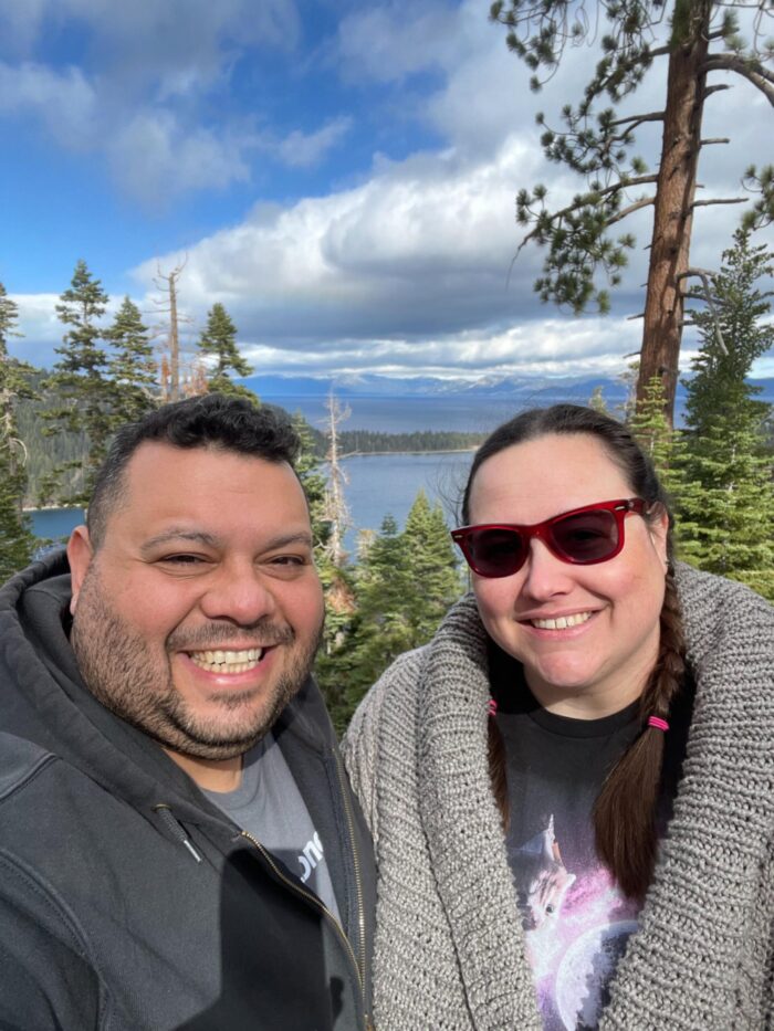 Lito and I smiling at the camera with a beautiful view in the background. There's a very blue lake, pine trees, and stormy clouds rolling in
