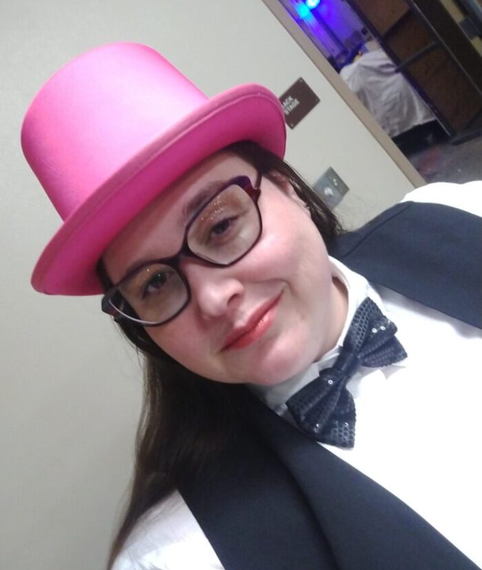 me ready for my jazz dance performance. I'm wearing a pink top hat, sparkly black bow tie, and a black vest over a white shirt. I have glittery gold eye makeup and red lipstick on.