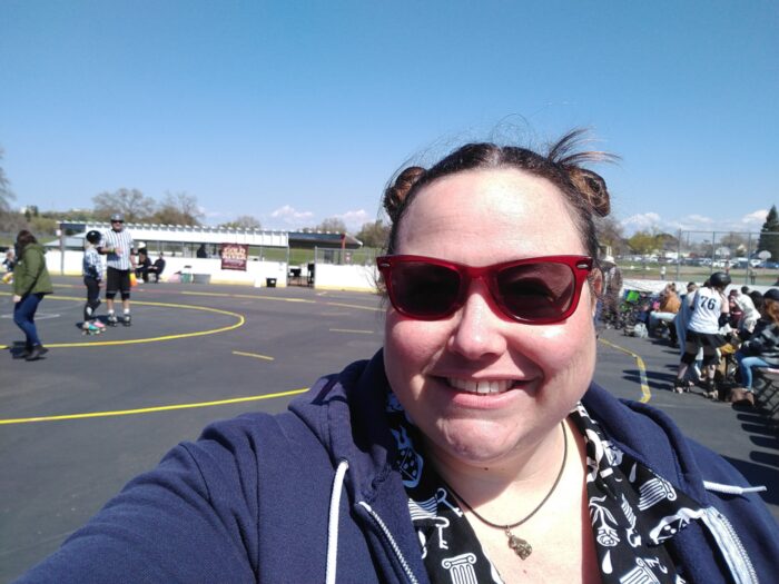 me, outside, wearing cherry-red sunglasses and smiling in front of a roller derby track. My hair is in two buns on top of my head.