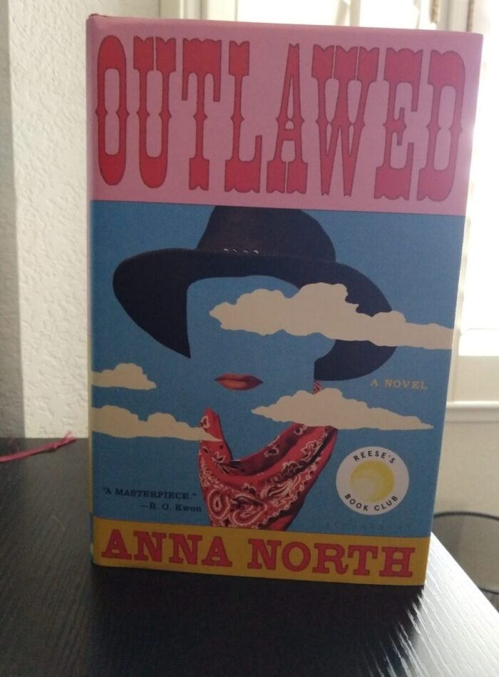 hardback book: Outlawed by Anna North. Cover features the suggestion of a face. There's a cowboy hat, red lips, and a bandana