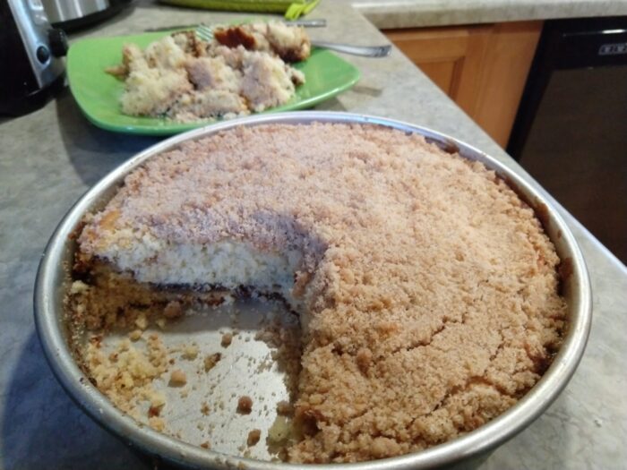 a nine-inch round pan of coffee cake with a piece missing. In the background, there is a plate with a completely crumbled up pile of coffee cake