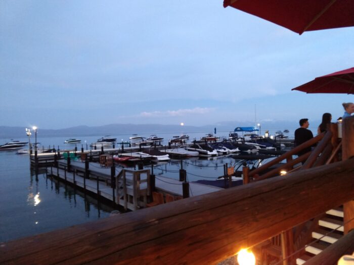 the moon coming up over the mountains in lake tahoe. Its reflection is visible on the water. Boats and a small marina are in the foreground