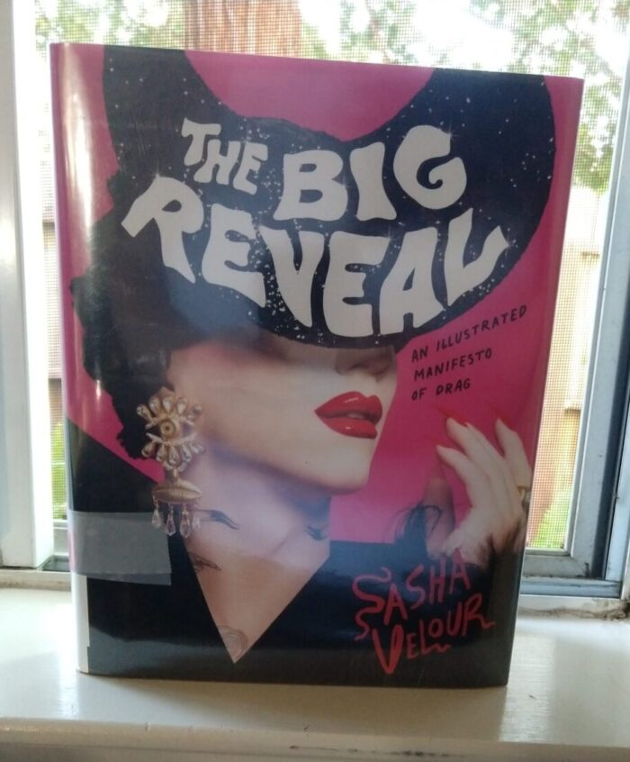 hardback book The Big Reveal. Cover features the author, Sasha Velour, in drag with shiny red lips and her face obscured by a huge fascinator