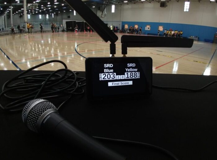 a tablet showing a score of SRD blue 203 points to SRD yellow 188 points. There's a microphone on the table in front of the tablet and a sports floor in the background