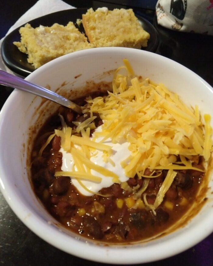 A bowl of homemade chili topped with cheese and sour cream, a plate of cornbread in the background