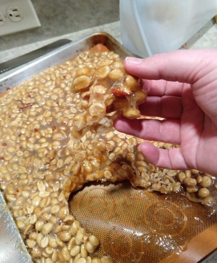 a handful of peanut caramel goop with a whole pan full of the same in the background