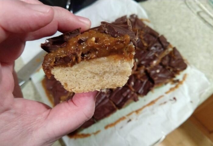 a piece of shortbread topped with dulce de leche caramel and a layer of chocolate held in hand. The rest of the batch is in the background