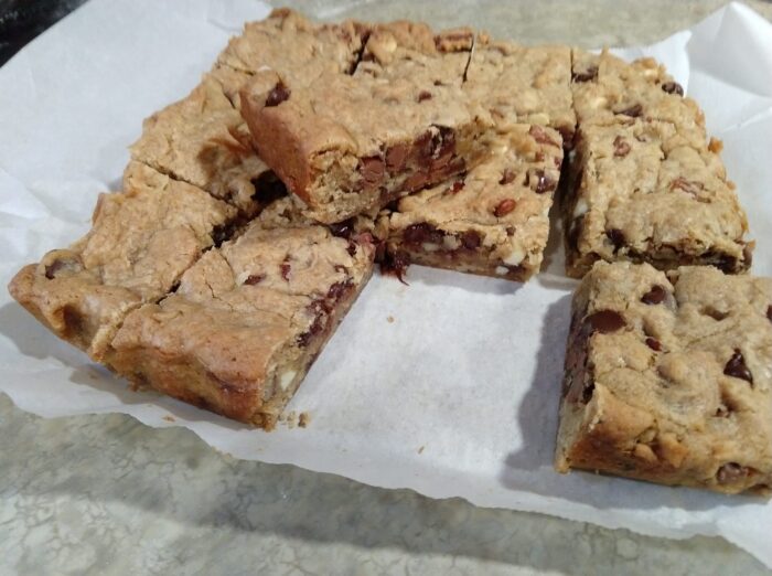 a sliced batch of chocolate chip cookie bars on top of a parchment paper. Bars are loaded with different kinds of chocolate chips, coconut, and pecans