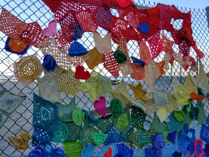 Knit and crochet squares and shapes zip tied to a chain-link fence with the ocean in the background. The yarn is roughly arranged by rainbow colors
