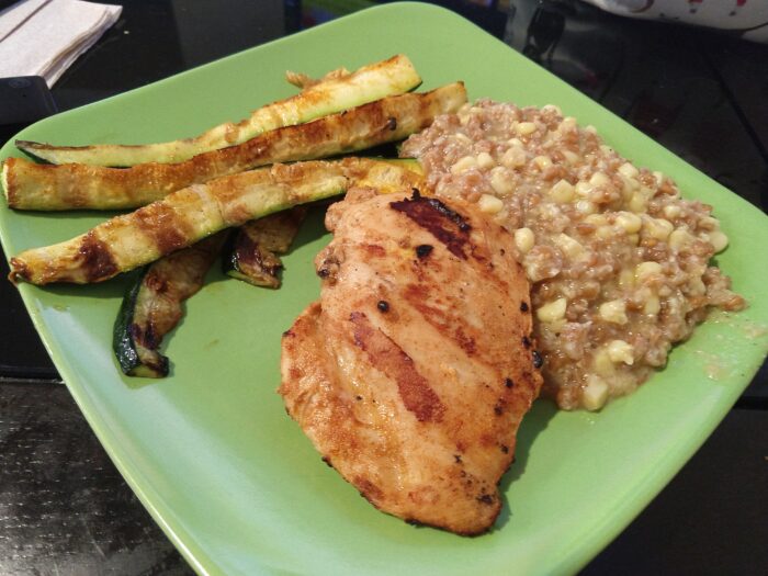 a plate of grilled chicken, grilled slices of zucchini, and buttered corn farro