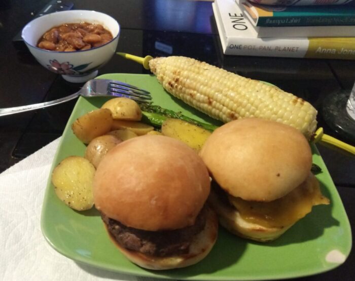 a meal of cheeseburger, roasted potatoes, asparagus, corn on the cob, and baked beans