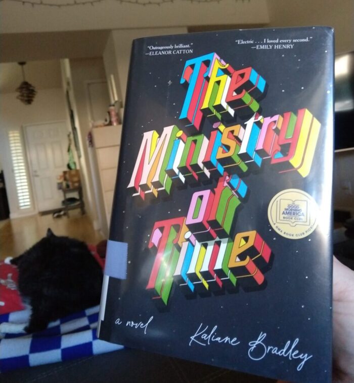 hardcover book: The Ministry of Time. Huey the cat is lying down on the couch in the background