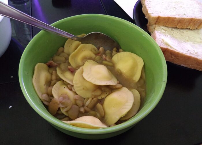 A bowl of brothy beans with pancetta and some ravioli mixed in, slices of bread on the side