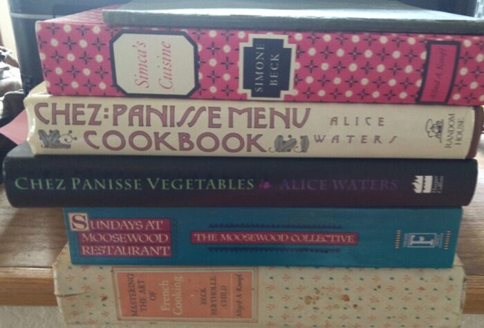 A pile of vintage cookbooks including Mastering the Art of French Cooking, Sundays at Moosewood Restaurant, and Chez Panisse Menu Cookbook